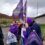 A group of people holding purple flags and signs stands outside on a cloudy day, engaging in conversation.