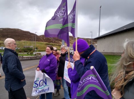 A group of people holding purple flags and signs stands outside on a cloudy day, engaging in conversation.