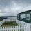 Small ferry terminal building by the sea, with a white fence in the foreground and boats moored in the background under a cloudy sky.