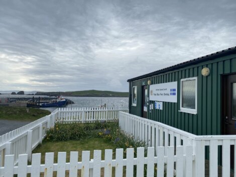 Small ferry terminal building by the sea, with a white fence in the foreground and boats moored in the background under a cloudy sky.
