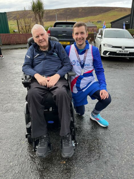 A man in a wheelchair and a man in athletic gear pose together outdoors. The athlete is kneeling beside the chair, smiling, with a medal around his neck.