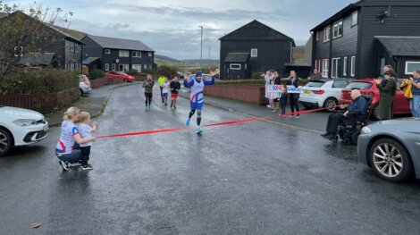 A runner crosses the finish line of a race in a neighborhood street, cheered by spectators. A woman and child hold the finish tape. Signs with supportive messages are visible in the crowd.