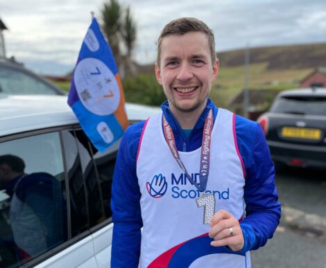 A man smiling, wearing a Mind Scotland bib, holding a medal with the number "1" on it. There's a Mind Scotland flag attached to a nearby car in the background.