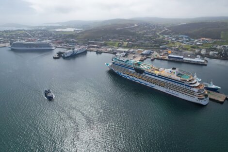 Aerial view of a harbor with three large cruise ships docked and a smaller boat navigating the water. Buildings and hills are visible in the background.