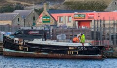 A boat docked at a harbor with two people working on it. Buildings and a truck labeled "Allanoff" are in the background.