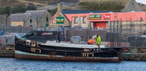 A boat docked at a harbor with two people working on it. Buildings and a truck labeled "Allanoff" are in the background.