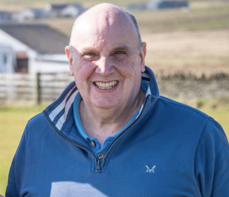 A man in a blue jacket smiles outdoors with blurred buildings and fields in the background.