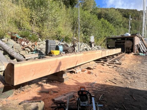 A long wooden beam is placed on supports outdoors, surrounded by tools and equipment, with trees and a wooden structure in the background.