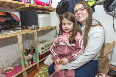 A woman and a young girl sit in a room with shelves containing pet supplies. A cat is visible in a cage on the shelf.