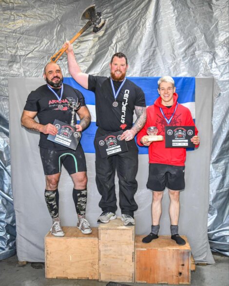 Three men stand on podiums holding plaques and medals; the central figure holds an axe aloft.