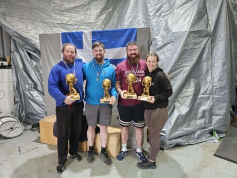 Four people stand in front of a flag, each holding a trophy shaped like a human skull. They are in a room with a metallic tarp backdrop.