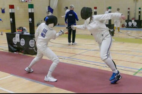 Two fencers in protective gear engaged in a match in a gymnasium, with one lunging toward the other.