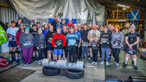 A group of people poses with awards in a gym, featuring weightlifting equipment in the foreground and a Scottish flag on the wall.