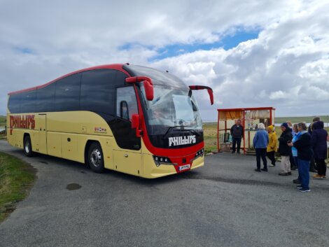 A red and yellow coach bus is parked on a road. A group of people are gathered nearby, looking at the bus. A small shelter is visible on the right under a cloudy sky.