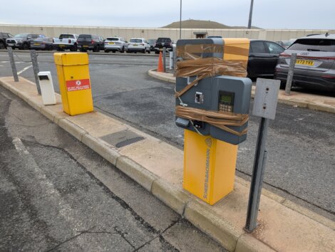 A parking ticket machine is wrapped with brown tape, next to a yellow waste bin. Several parked cars are in the background.