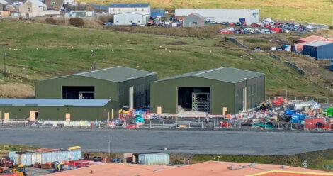 Two green industrial warehouses are under construction, surrounded by building materials and machinery on a hillside with additional buildings and parked cars in the background.