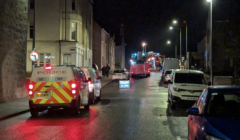 Night scene on a street with police and fire vehicles parked, lights flashing. A "Police Slow" sign is visible in front of a police van.