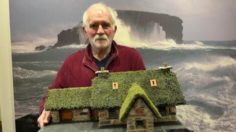 A man holding a model of a stone house with a grass-covered roof; a large ocean wave is shown in the background.