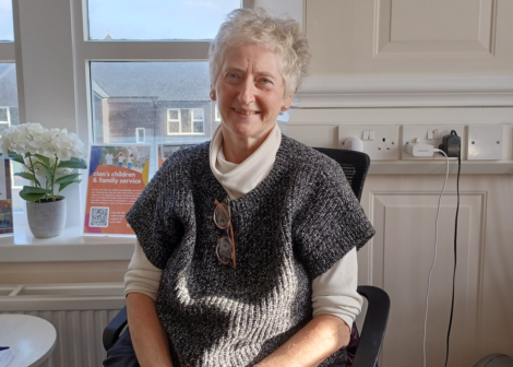 An elderly person with white hair sits in an office chair, wearing a gray knitted vest over a cream turtleneck. A potted plant and sign are on the windowsill in the background.