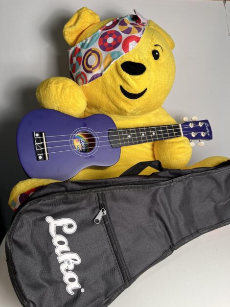 A yellow teddy bear wearing a colorful bandana holds a purple ukulele. In front is a black ukulele case with "Laka" written on it.