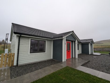 Modern single-story house with gray siding, a red front door, and a gabled roof. Surrounded by a lawn and a gravel driveway on a cloudy day.