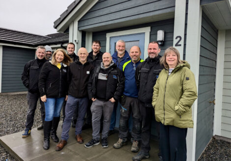 A group of eleven people stand smiling in front of a gray house, wearing casual and outdoor clothing, on a cloudy day.