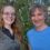Two people are standing outdoors in front of greenery, smiling at the camera. The person on the left has long red hair and glasses, and the person on the right has short gray hair and is wearing a blue shirt.