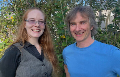 Two people are standing outdoors in front of greenery, smiling at the camera. The person on the left has long red hair and glasses, and the person on the right has short gray hair and is wearing a blue shirt.