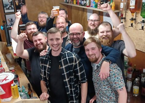 A group of men in casual clothing gather at a bar, smiling and raising their fists in celebration. Bottles and drinks are visible in the background.