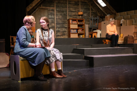 Two women sit and converse on stage, while another woman sits in the background. The set resembles a cozy room with stairs and shelves.