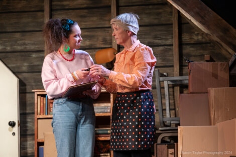 Two people stand in a room with cardboard boxes. One is holding a clipboard, and the other is gesturing with her hands while wearing a headscarf and apron.