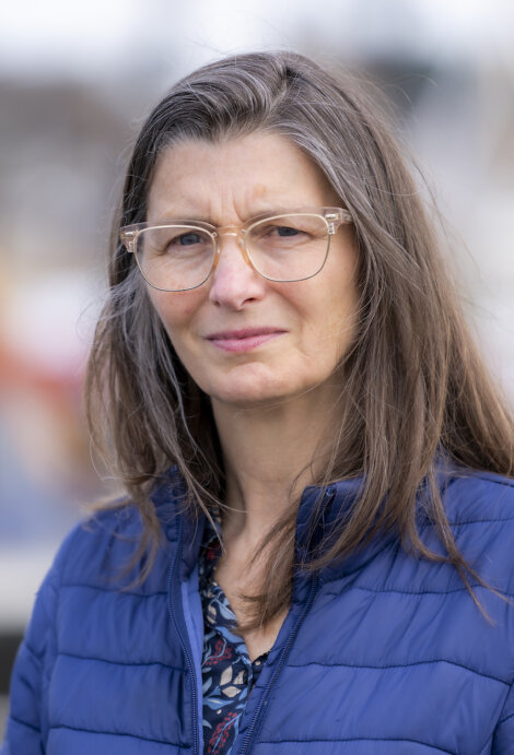 A woman with long hair and glasses is wearing a blue jacket and standing outdoors.