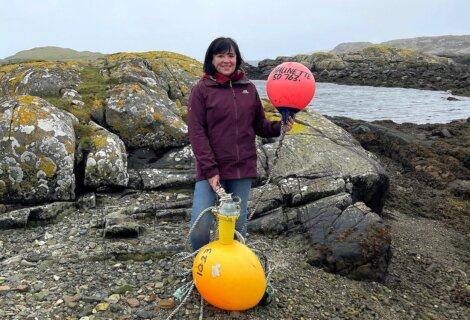 A woman stands on a rocky shore holding two buoys, one orange and marked "1023" and the other red and marked "SO 763." A small body of water is visible in the background.