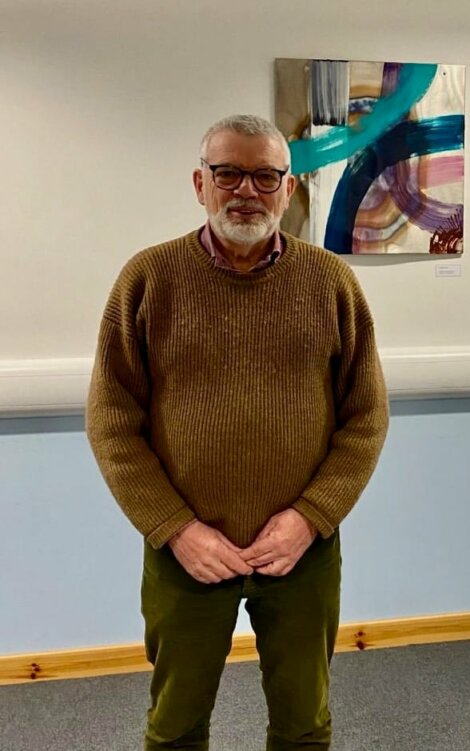 An older man with glasses and a beard stands indoors wearing a brown sweater and green pants. A colorful abstract painting is on the wall behind him.