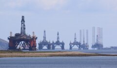 View of offshore oil drilling platforms lined up near the coast, with a body of water in the foreground.
