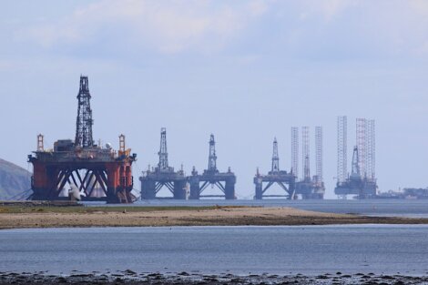 View of offshore oil drilling platforms lined up near the coast, with a body of water in the foreground.