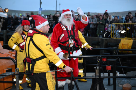 A person dressed as Santa Claus waves from a boat alongside RNLI crew members in yellow uniforms. A crowd of onlookers stands in the background.