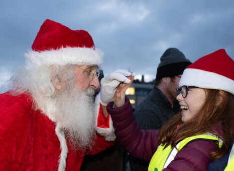 A person dressed as Santa Claus interacts with a smiling girl wearing glasses and a Santa hat. Both are outside, and the sky is overcast.