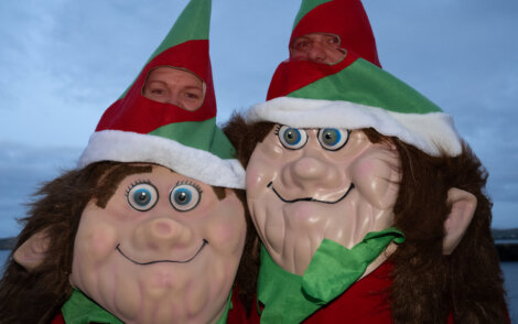 Three people dressed as Christmas elves, with two wearing large masks, pose outdoors against a cloudy sky.
