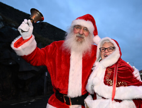 Two people in Santa outfits smile, one holding a bell, outdoors with a rocky background.