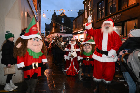 Santa and elves parade through a festive street with onlookers and holiday decorations.
