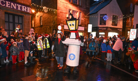 A festive street scene with a crowd, including people in Santa hats and high-visibility vests. A person is dressed as a lighthouse mascot in the center, with holiday lights overhead.