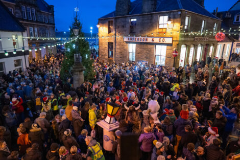 A large crowd gathers in a festive town square at dusk, surrounded by buildings and holiday lights.