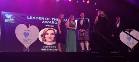 Group of people on stage at an award ceremony, with a screen displaying "Leader of the Year Award" and "Lucy Flaws, NHS Shetland.