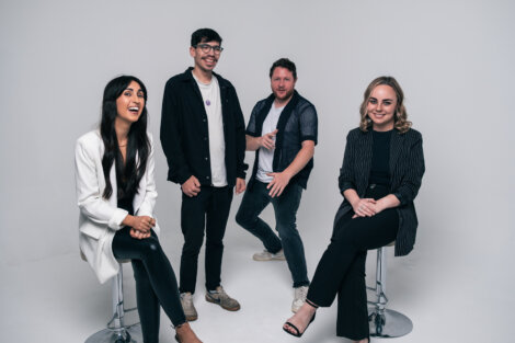 Four people pose together in a studio. Two are seated on stools, while the other two stand. The background is plain white.