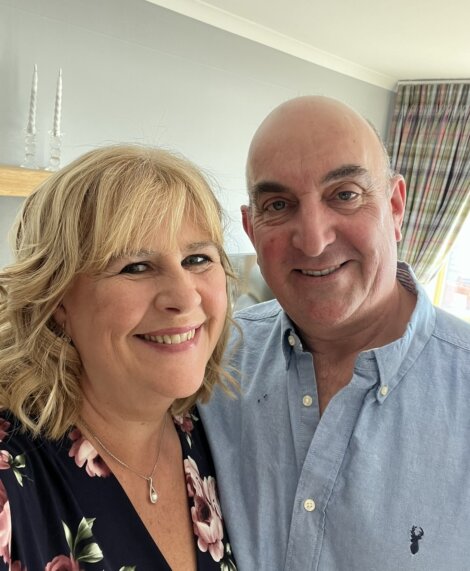 A man and woman smiling, standing close together indoors. The woman has blonde hair and wears a floral top, while the man is bald and wears a light blue shirt.