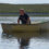 A man in a small rowboat interacts with a seal in the water. A grassy landscape is visible in the background.
