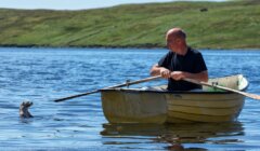 A man in a rowboat looks at an otter swimming nearby in a calm lake with grassy hills in the background.