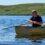 A man in a rowboat looks at an otter swimming nearby in a calm lake with grassy hills in the background.