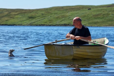 A man in a rowboat looks at an otter swimming nearby in a calm lake with grassy hills in the background.